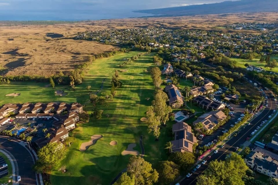 Drone shot of lush green fairways, surrounding homes, and scenic terrain rolling toward the ocean.

