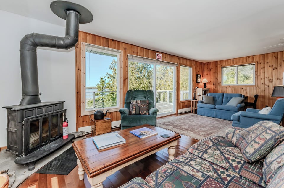 Cozy lakeside living room with wood-burning stove, rustic charm, and large windows offering stunning water views — the perfect spot to relax after a day of beach adventures!