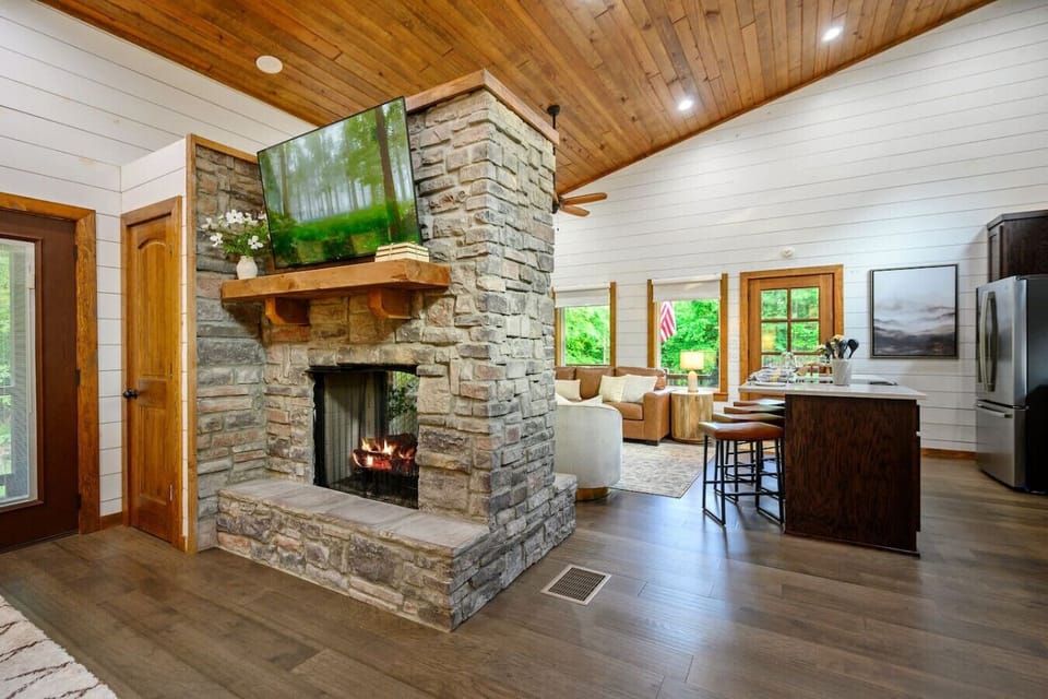 This open-concept living room is where the magic happens—fireplace, kitchen, dining, and those massive forest-facing windows all flowing together so seamlessly that it genuinely feels like the whole cabin is one big cozy hangout.