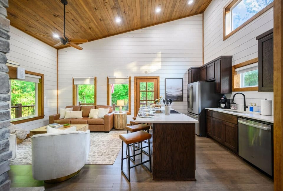 The whole vibe of this place just *works*—white shiplap walls, warm wood ceilings, a kitchen island that begs for conversation, and those floor-to-ceiling windows bringing the forest right into your gathering space.
