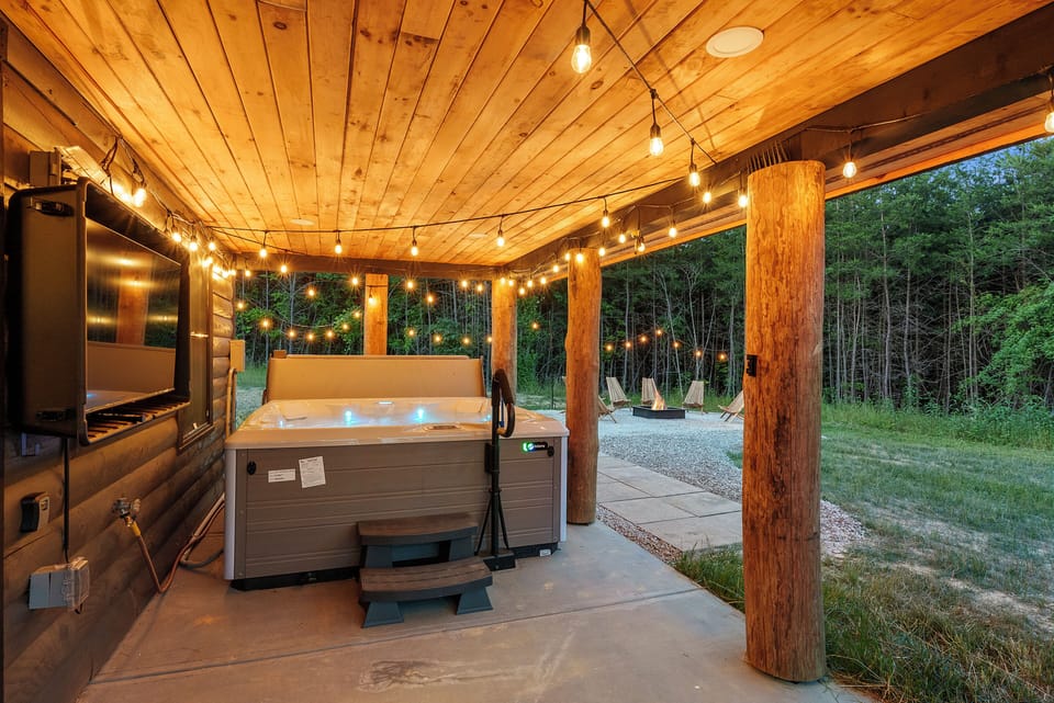 Relax in a private hot tub under string lights, with cozy covered patio and fire pit just steps away, perfect for peaceful nights in nature.