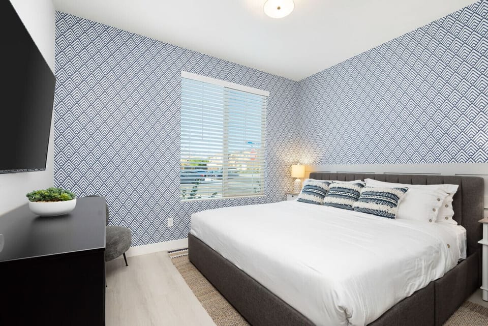King bedroom featuring bold modern wallpaper, soft white bedding, flat-screen TV, and plenty of natural light—perfect for relaxing after a pool day.