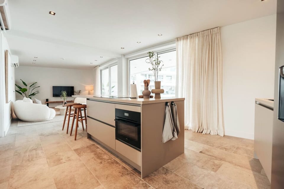 A spacious, airy kitchen island seamlessly flows into a cozy living area. Large windows invite natural light, highlighting the modern design and creating a welcoming atmosphere.