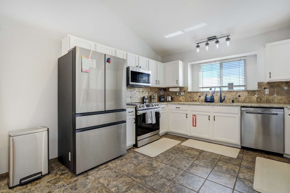 Bright kitchen with stainless steel fridge, oven, and dishwasher, white cabinets, and tiled floors. A window above the counter brings in daylight while you prep meals, making it easy to cook, pour a drink, and gather around the space.