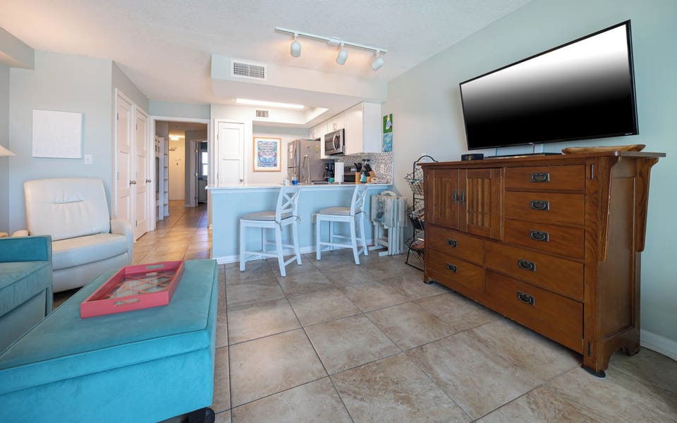 The living area features a large TV, stylish turquoise seating, and a view of the adjacent kitchen with modern appliances