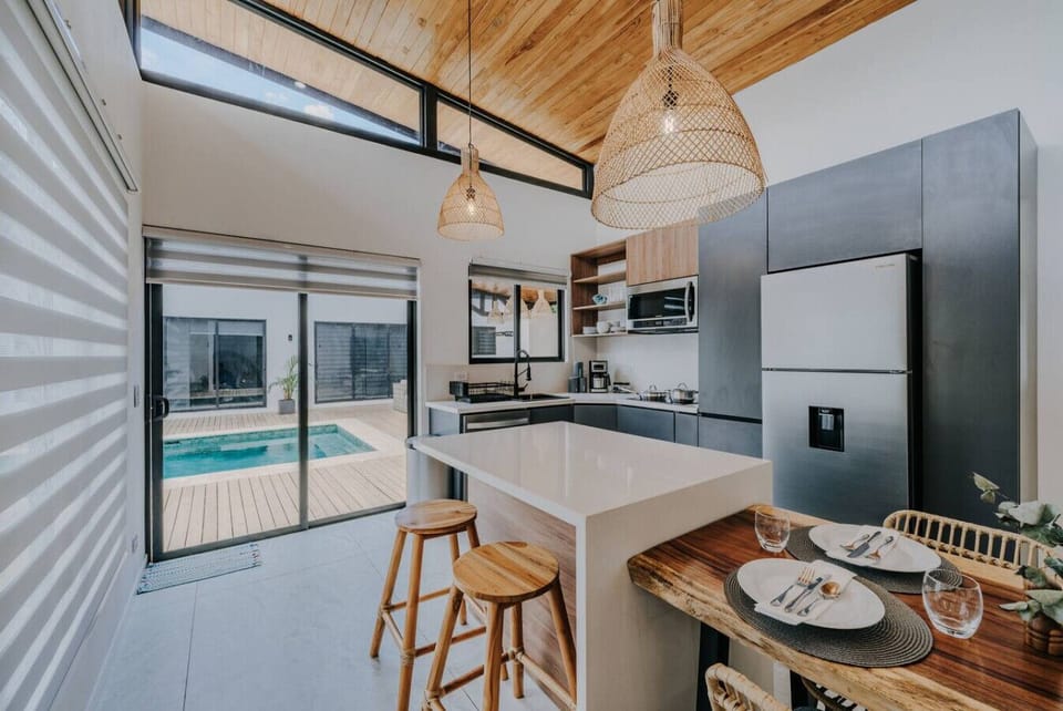 A beautifully designed modern kitchen with a bar counter lit by wicker-style pendant lights, two bar stools, and a view of the pool outside. The bar flows seamlessly into the dining area, perfect for both casual meals and entertaining.