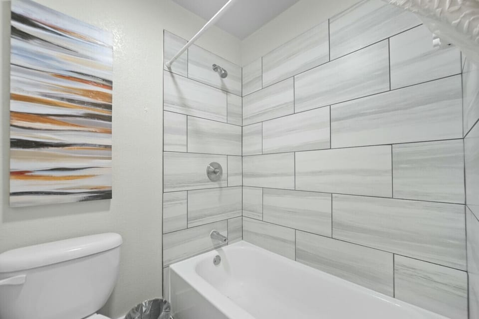 Clean bathroom interior highlighting bathtub shower enclosure with marbled grey tile, shower rod, toilet unit, and abstract canvas art.