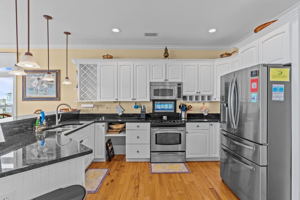 Oak Island NC vacation rental kitchen with white cabinets, black granite countertops, stainless steel refrigerator, stove, and microwave, wood floors, pendant lighting, double sink, and coastal artwork in a bright, open layout.