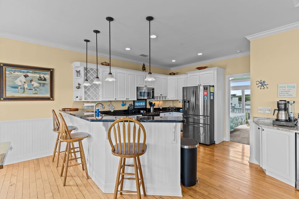 Oak Island NC vacation rental kitchen with white cabinets, black granite countertops, stainless steel appliances, pendant lighting, breakfast bar with stools, wood floors, coastal artwork, and open layout connecting to bedrooms.