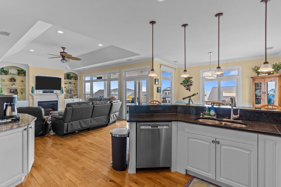 Oak Island NC vacation rental open-concept kitchen and living area with granite countertops, stainless dishwasher and sink, pendant lighting, wood floors, leather seating, fireplace, built-in shelves, and large windows leading to a deck.