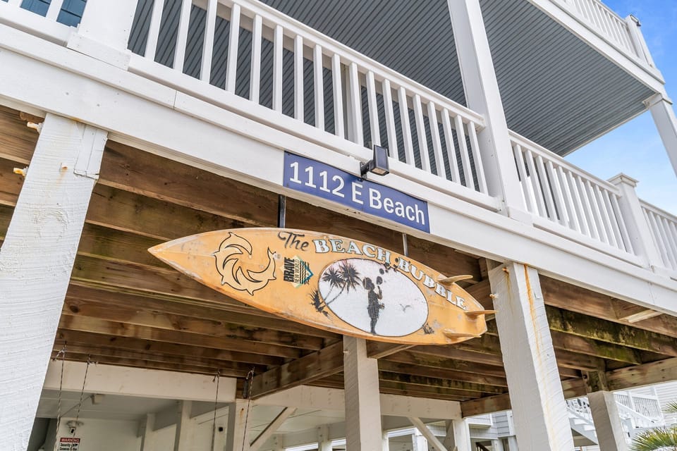 Close-up of an Oak Island NC vacation rental exterior showing the address sign “1112 E Beach,” white porch railings, elevated beach house supports, and a decorative surfboard sign reading “The Beach Bubble” beneath the home.