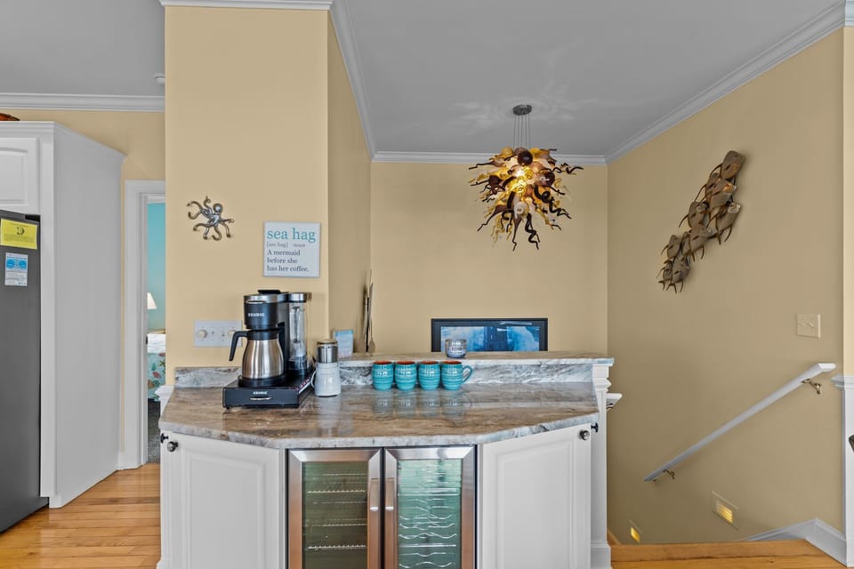 Oak Island NC vacation rental beverage station with granite countertop, wine fridge, coffee maker, mugs, coastal wall decor, pendant lighting, stairway access, and open connection to the kitchen and living area.