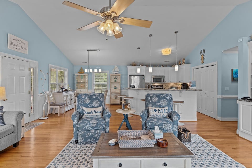 Bright open living space with vaulted ceilings, coastal decor, seating area, dining nook, and fully equipped kitchen with bar seating and modern appliances.