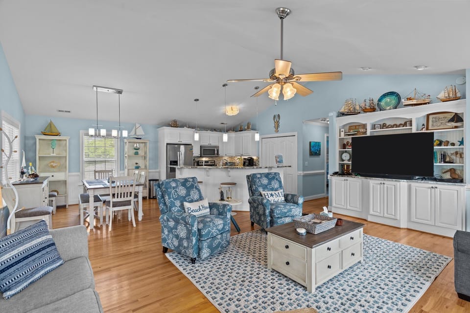 Bright open living space with vaulted ceilings, coastal decor, seating area, dining nook, and fully equipped kitchen with bar seating and modern appliances.