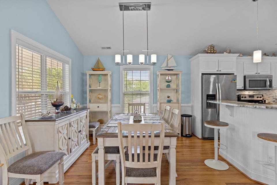 Bright dining space with table for four, coastal decor, large windows, decorative shelving, and access to outdoor deck, perfect for meals and gatherings.
