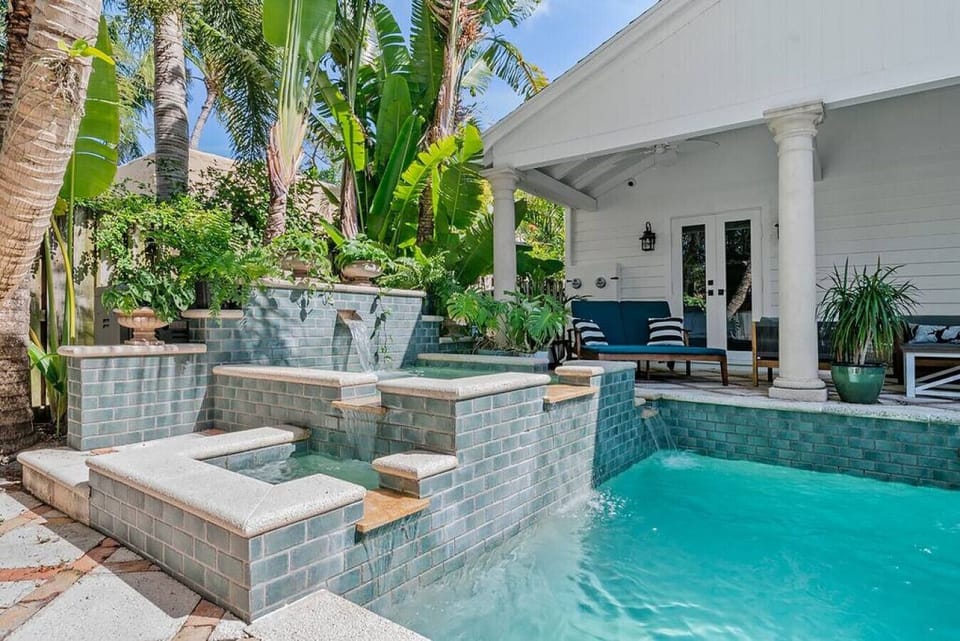 Daytime pool with tiled waterfall feature, cascading spillway, tropical palms, and columned covered lounge seating alongside