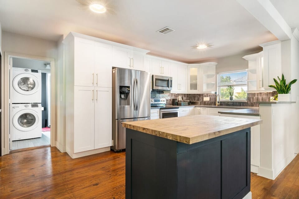 Bright and modern kitchen features white cabinetry, stainless steel appliances, and a contrasting dark kitchen island with a wooden countertop.

