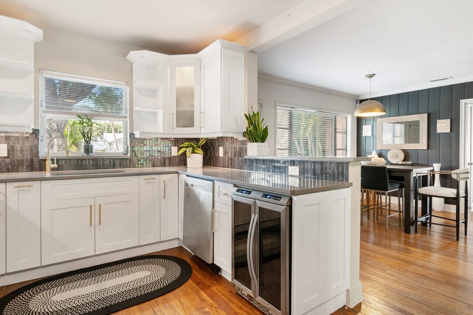 Modern kitchen boasts sleek white cabinetry, stainless steel appliances, and a view into a bright dining area with a dark table.
