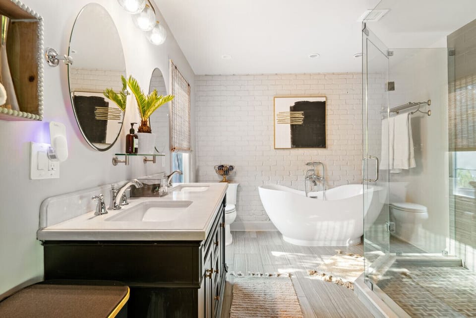 Sunlit primary bath with freestanding soaking tub, glass-enclosed shower, oval vanity mirror, and white-painted brick walls