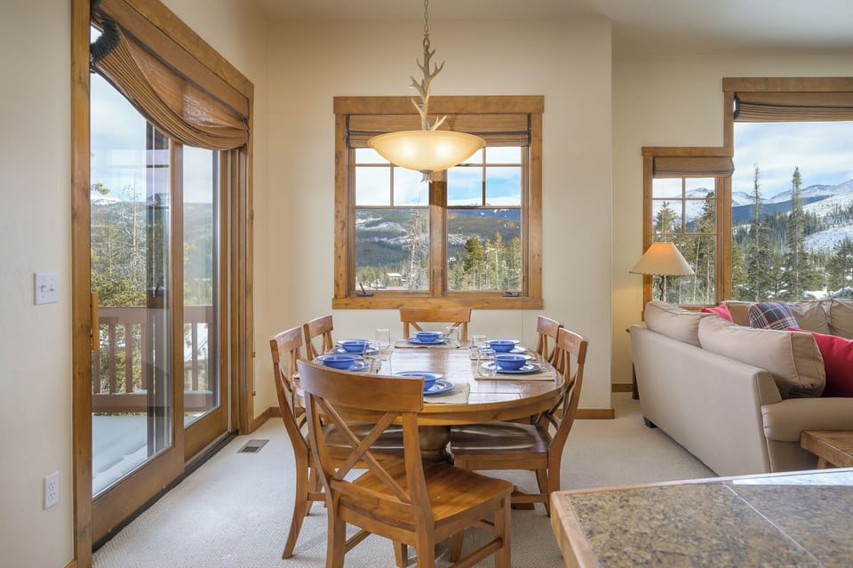 - Dining area with round wooden table set for six overlooks scenic mountain views   - Large windows and sliding glass doors brighten the space with natural light   - Cozy setup makes it perfect for family meals in an inviting alpine atmosphere