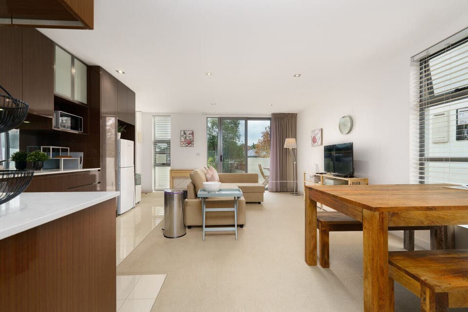 A versatile living space with a substantial wooden dining table, a modern TV setup, and large windows that bring in natural light.