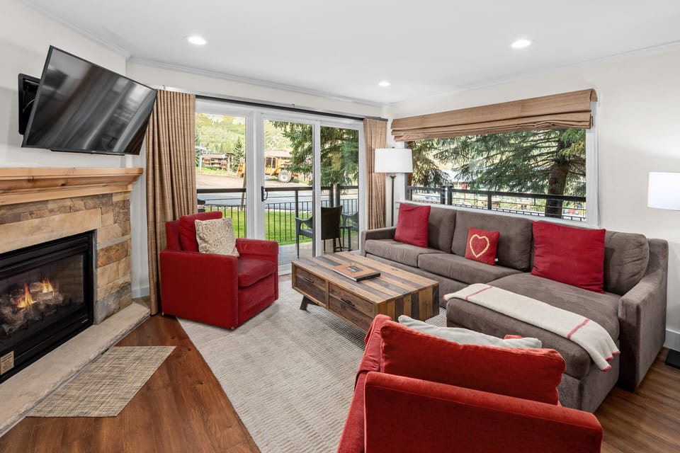 Cozy living area featuring a plush sectional sofa, stone fireplace with mounted TV, and expansive windows that open to a private patio.