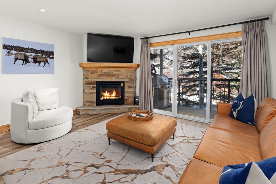 Cozy living area featuring a leather sectional sofa, stone fireplace with mounted TV, and expansive windows that open to a private balcony.