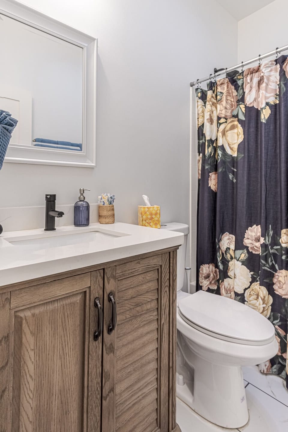 Full Bathroom: Refresh with ease in this stylish full bathroom, equipped with a rustic wood vanity, sleek white countertop, modern black matte fixtures, bright mirror lighting, and a full shower and tub combo accenting a floral curtain.