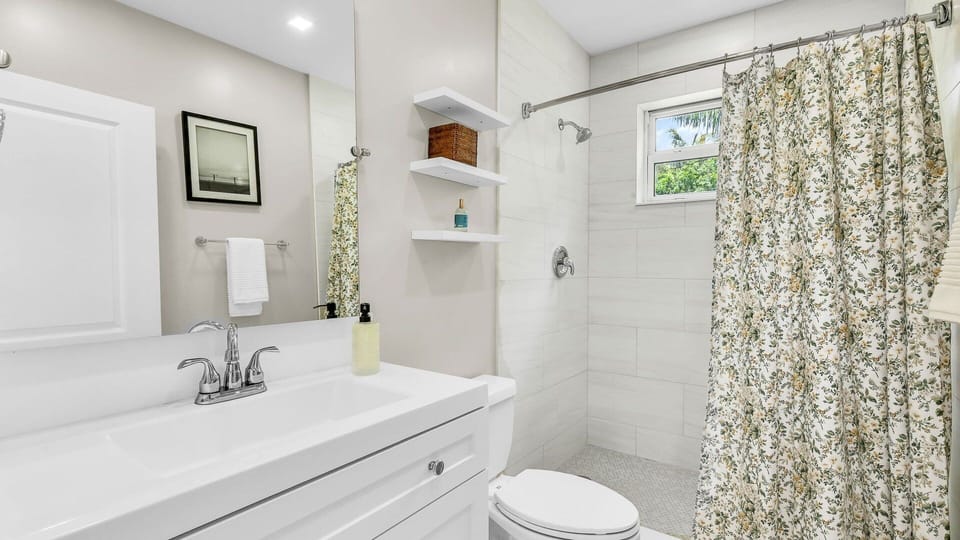 This shared bathroom between Bedrooms 1 and 2 features a clean, modern design with a combination tub and shower, ideal for both quick rinses and relaxing soaks. Thoughtfully stocked with essentials and open shelving for added convenience.