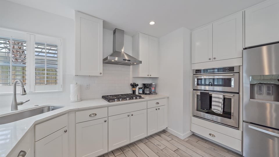This fully updated gourmet kitchen features sleek, white coastal cabinetry, a subway tile backsplash, a gas cooktop, and built-in double ovens perfect for any vacation chef.