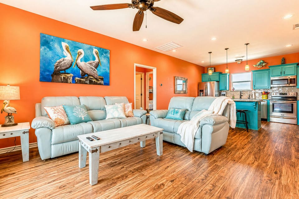 Open-concept living area with plush sofas, wood flooring, ceiling fan, and seamless flow into the kitchen space