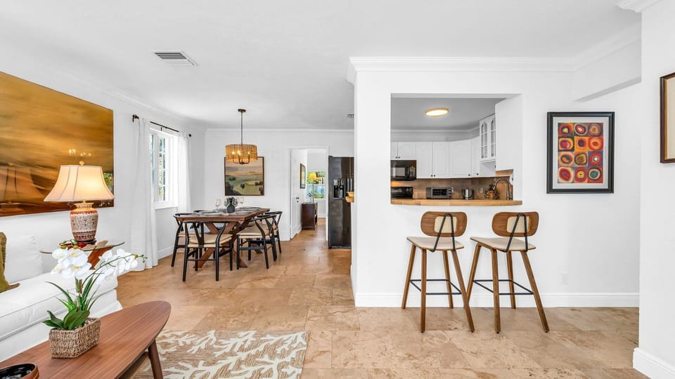 This stylish dining area comfortably seats six and is perfect for family meals or casual entertaining. Framed by natural light from the garden-view window, it features a modern wood table, woven pendant lighting, and tropical-inspired dinnerware for a touc
