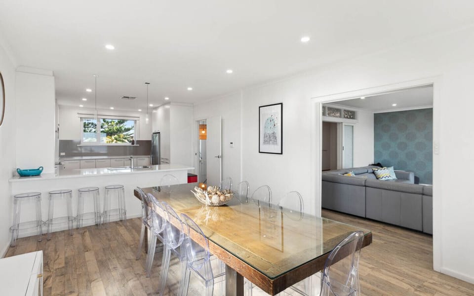 A modern, open-plan living space with a sleek design. It features a large dining table with transparent chairs in the foreground, a white kitchen area with bar stools, and a cozy living room with a gray sofa and patterned wallpaper in the background. The 