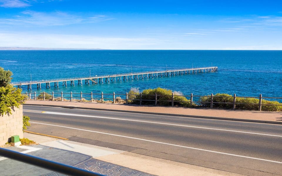 A scenic view of a road with a sidewalk and a fence beside it, leading towards a long pier extending into a clear blue body of water under a bright sky.