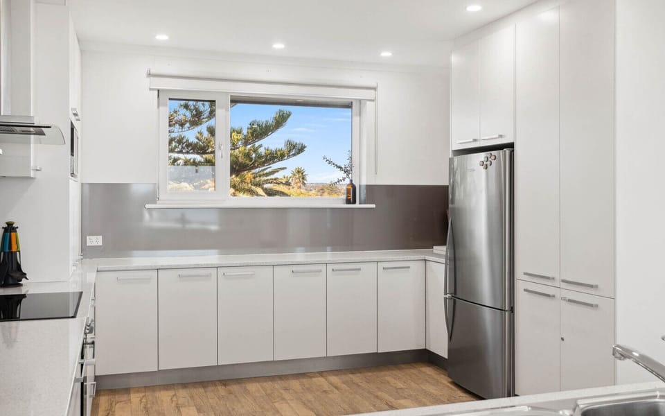 A modern kitchen with a clean and bright design. It features white cabinetry, a stainless steel refrigerator, and a window that offers a view of trees outside, creating a fresh and airy atmosphere.