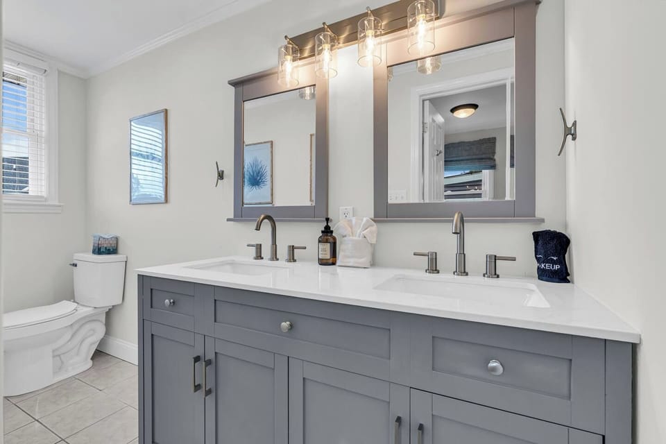 Full bath with double-vanity gray cabinet, white quartz top, two framed mirrors, and brass fixtures.