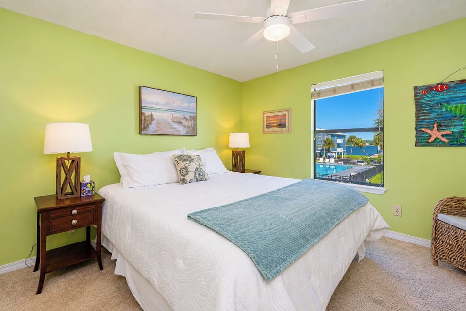 The master bedroom features a large bed, vibrant green walls, and a lovely view of the pool area and palm trees outside