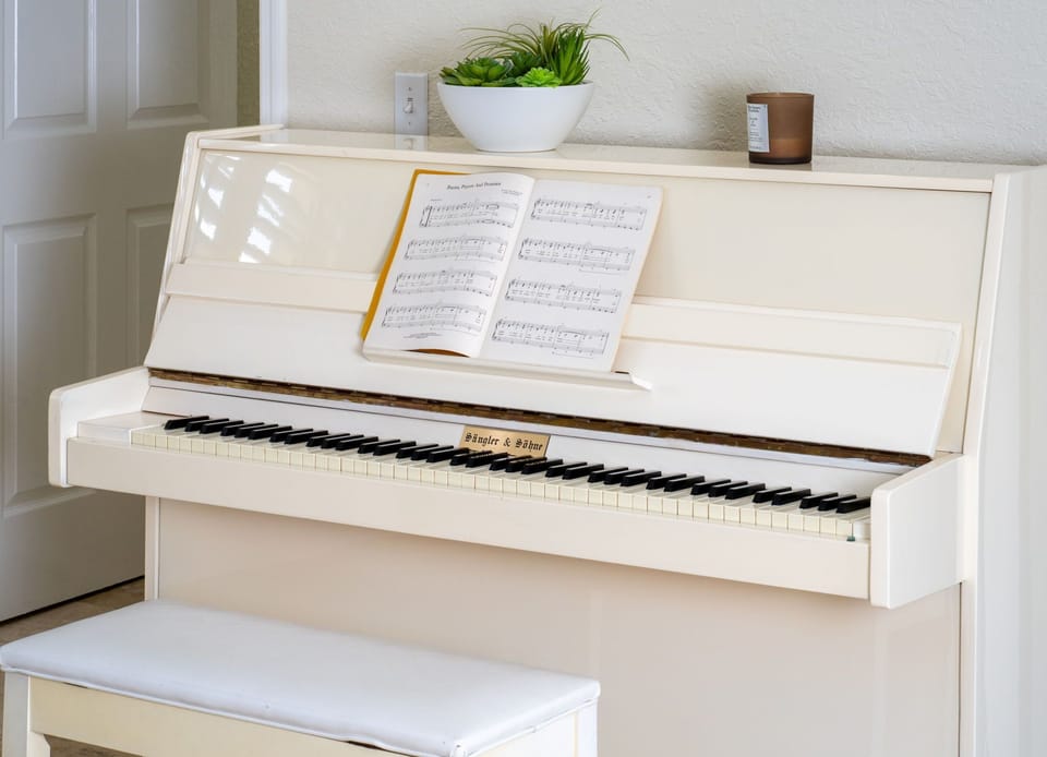 A sleek ivory upright piano with an open music book, topped with a potted plant and candle for a touch of charm.