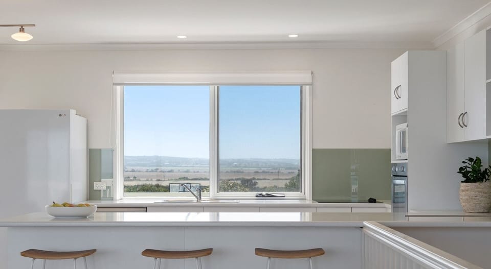 A modern kitchen with white cabinetry and a large window offering a view of the landscape. There are three stools at the counter, a bowl of fruit on the counter, and a potted plant adding some greenery to the space.