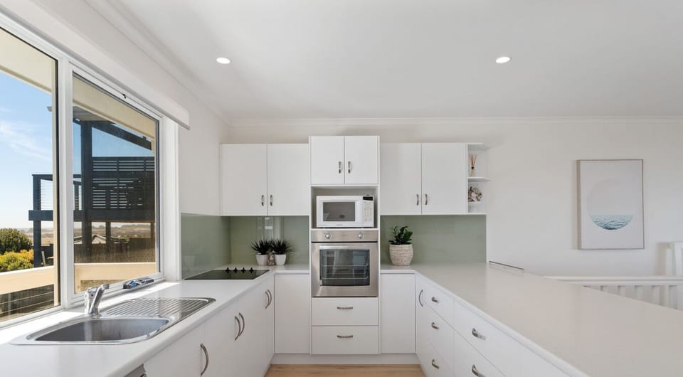 A modern, bright kitchen with predominantly white cabinetry and countertops. It features a stainless steel sink, an oven, a microwave, and a dishwasher. The kitchen has a light wooden floor and large windows that allow natural light to flood in. There are