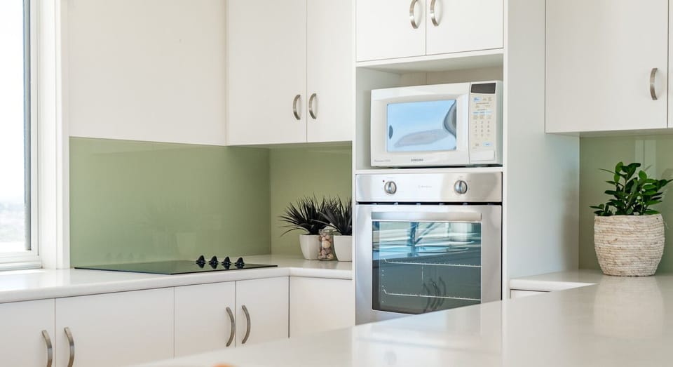 A modern kitchen with white cabinets and a light green backsplash. It features a stainless steel oven, a microwave placed above it, and a bowl of apples on the countertop. A potted plant adds a touch of greenery to the clean and stylish space.