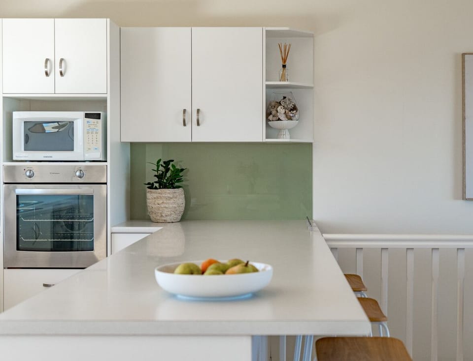 A modern kitchen area with white cabinetry, a stainless steel oven, and a microwave. There is a green backsplash and a white countertop with a bowl of fruit placed on it. The kitchen also features a decorative plant and a framed artwork on the wall, givin