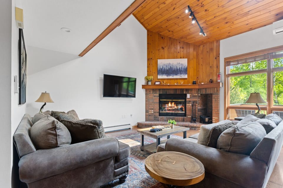 Open-concept living with forest views, deck access, cozy fireplace, and plush couches—perfect for relaxing or gathering together.
