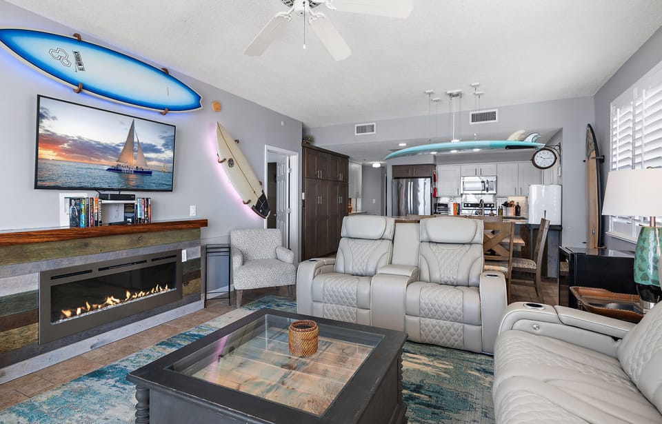 The Gulf View Living Room features a large Smart TV, electric fireplace, backlit surfboard decor, and brand new, leather reclining sofa and loveseat!