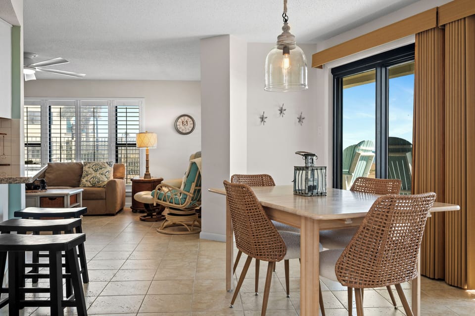 Step into your bright, airy dining space where woven chairs invite lingering conversations around the modern table, with ocean breezes flowing through sliding doors.