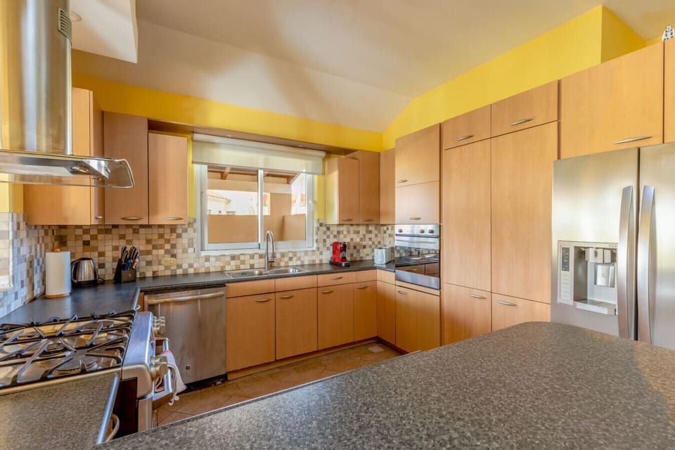 Modern kitchen featuring wood cabinetry, sleek countertops, and stainless steel appliances. Includes a gas stove, oven, dishwasher, and a spacious fridge, all complemented by a stylish mosaic tile backsplash and natural light from a large window.
