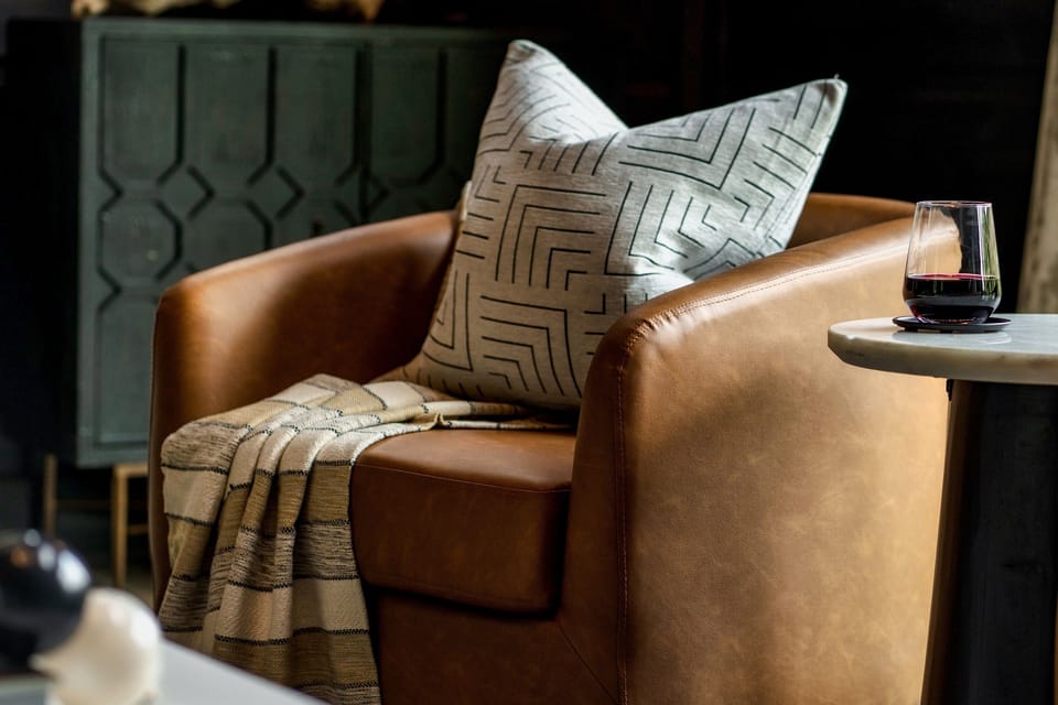 This cozy corner offers a perfect spot for quiet moments, featuring a luxurious brown leather armchair adorned with a modern geometric pillow and a soft plaid throw. A nearby marble-topped side table holds a glass of red wine, inviting you to sit back and