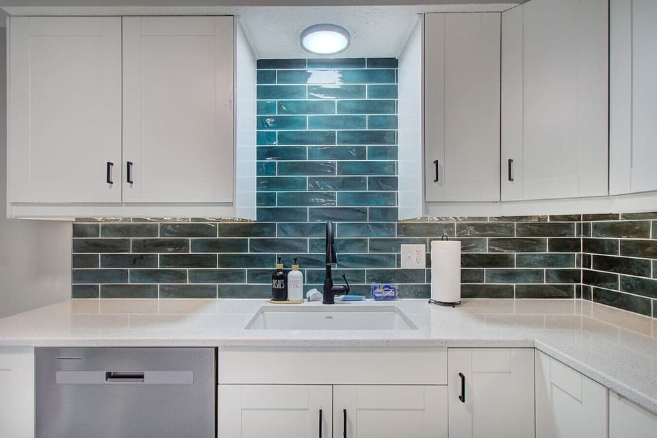 Kitchen sink area with white cabinetry, dishwasher, and a colorful tile backsplash.