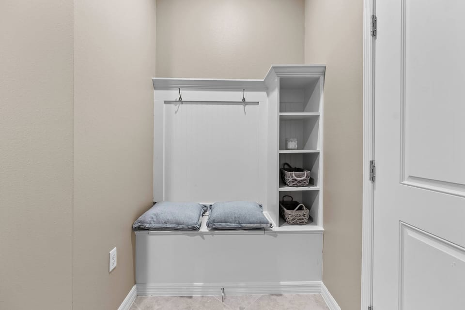 Welcome to our inviting mudroom, designed for comfort with a cozy built-in bench and soft gray cushions, ideal for unwinding after your adventures. The light beige walls and stylish storage baskets create a warm, organized space.