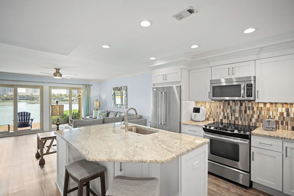 Open-concept kitchen with granite island, stainless steel appliances, and modern backsplash. Flows into a bright living area with lake views and stylish, coastal decor.